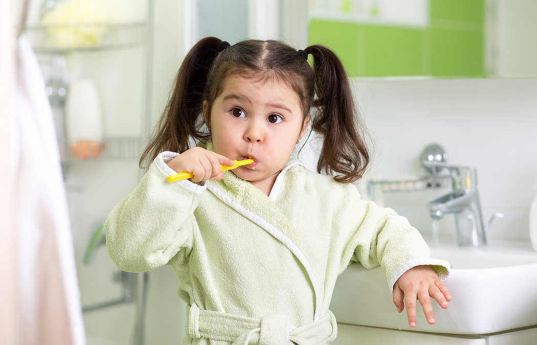 A young child with pigtails is brushing their teeth in a bathroom, wearing a green robe and holding a yellow toothbrush., 