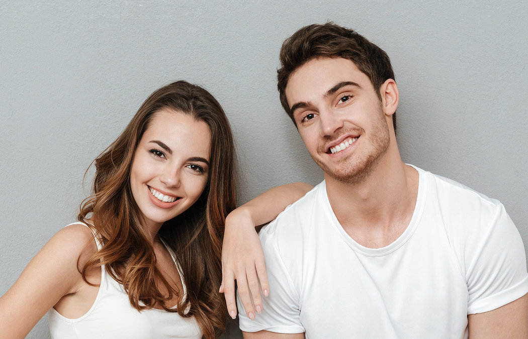 A smiling man and woman in white shirts stand against a gray background, with the woman's arm resting on the man's shoulder., 