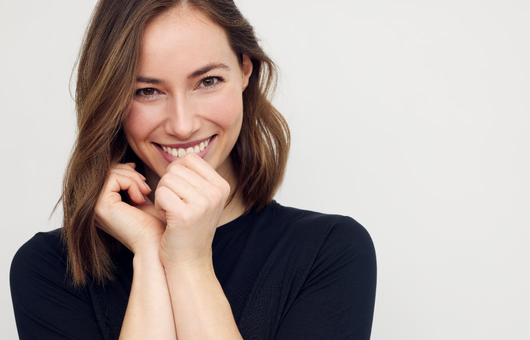 Woman with shoulder-length hair smiling and holding her hands near her face, wearing a black top against a plain background., 