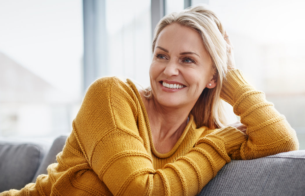 A woman in a yellow sweater sits on a couch, smiling and looking to her left with her hand resting on her head., 