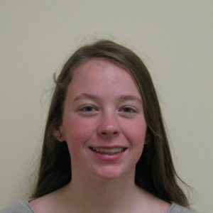 A young person with long brown hair smiles at the camera against a plain light-colored background.
