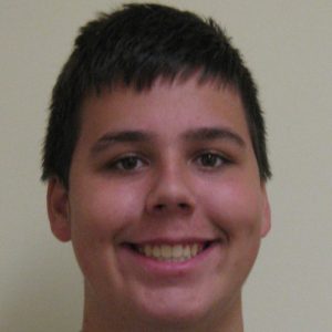 A young person with short dark hair is smiling at the camera against a plain light-colored background.