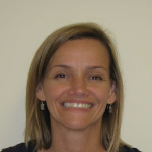 A woman with straight, shoulder-length blonde hair is smiling at the camera against a plain light-colored background.