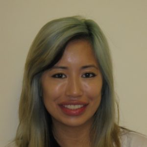 A woman with long, light greenish-blonde hair is smiling at the camera against a plain light-colored background.