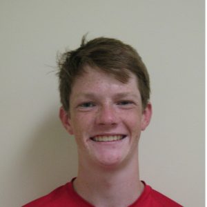 A young person with fair skin, short brown hair, and freckles smiles at the camera while wearing a red shirt against a plain light-colored background.