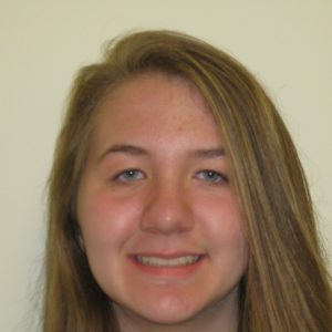 A young person with long, straight, light brown hair is smiling slightly in front of a plain, light-colored background.