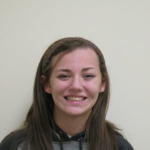 A young woman with straight brown hair, wearing a dark hoodie and headband, smiles at the camera against a plain light-colored background.