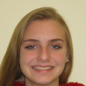 A young person with long blonde hair smiles at the camera against a plain light-colored background.