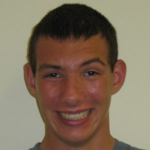 A young person with short dark hair smiles broadly at the camera against a plain light-colored background.