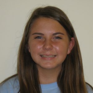 A young woman with long brown hair smiles in front of a plain, light-colored background. She is wearing a light blue shirt.