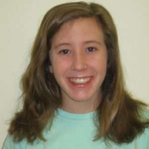 A young person with long, light brown hair and a light blue shirt smiles in front of a plain, light-colored background.