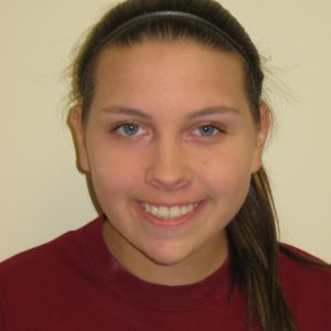A young person with long brown hair pulled back, wearing a maroon shirt, is smiling at the camera against a plain light-colored background.