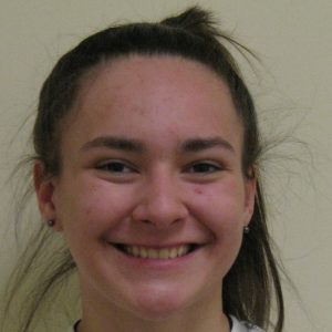 A young woman with long brown hair in a ponytail is smiling at the camera against a plain light-colored background.