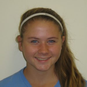 Teen girl with long blonde hair, wearing a white headband and blue shirt, smiling at the camera against a plain light-colored background.