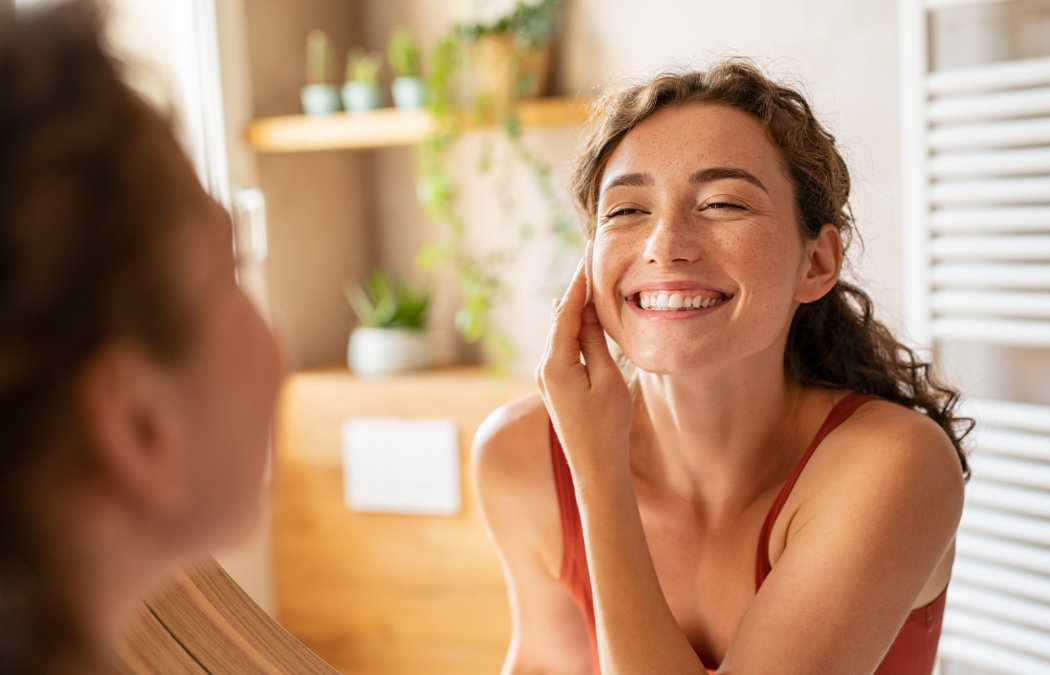 Woman smiling and applying skincare cream to her cheek while looking in the mirror in a bright bathroom with plants in the background., 