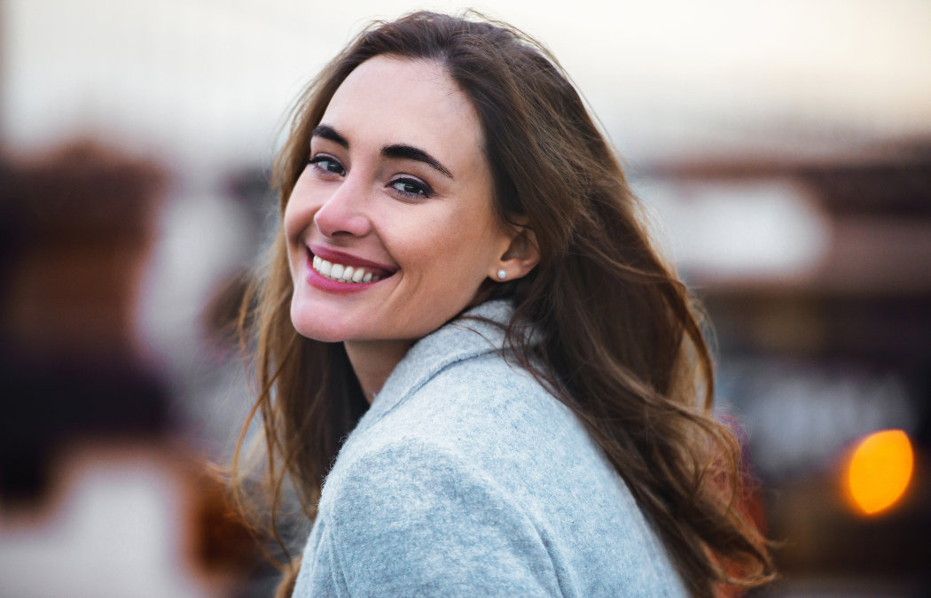 A woman with long brown hair and a light gray coat smiles at the camera outdoors, with a blurred background., 