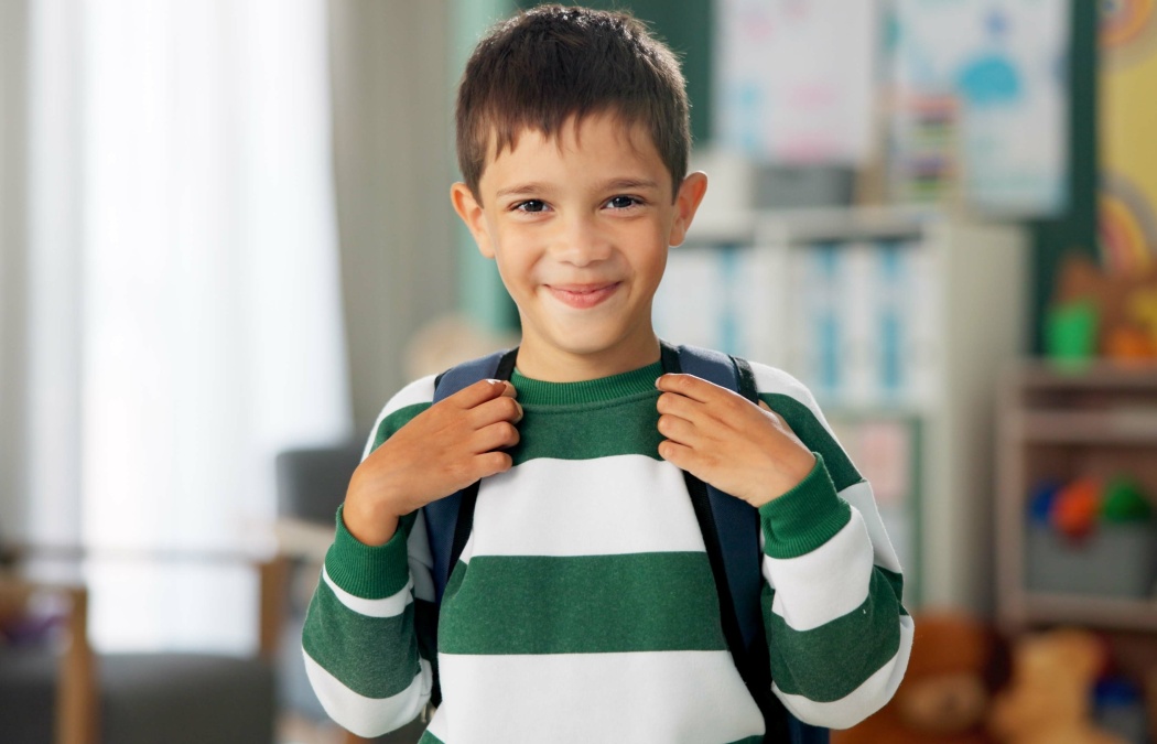 Child, face and laughing in school classroom or education with backpack for kindergarten, learning or joke. Boy, kid and portrait for study development on academy campus or student, humor or funny, 