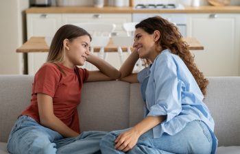 Loving mother listening to daughter with empathy and understanding while sitting together on sofa bonding at home, pre-teen girl child sharing secrets with mom, parent communicating with teenager
